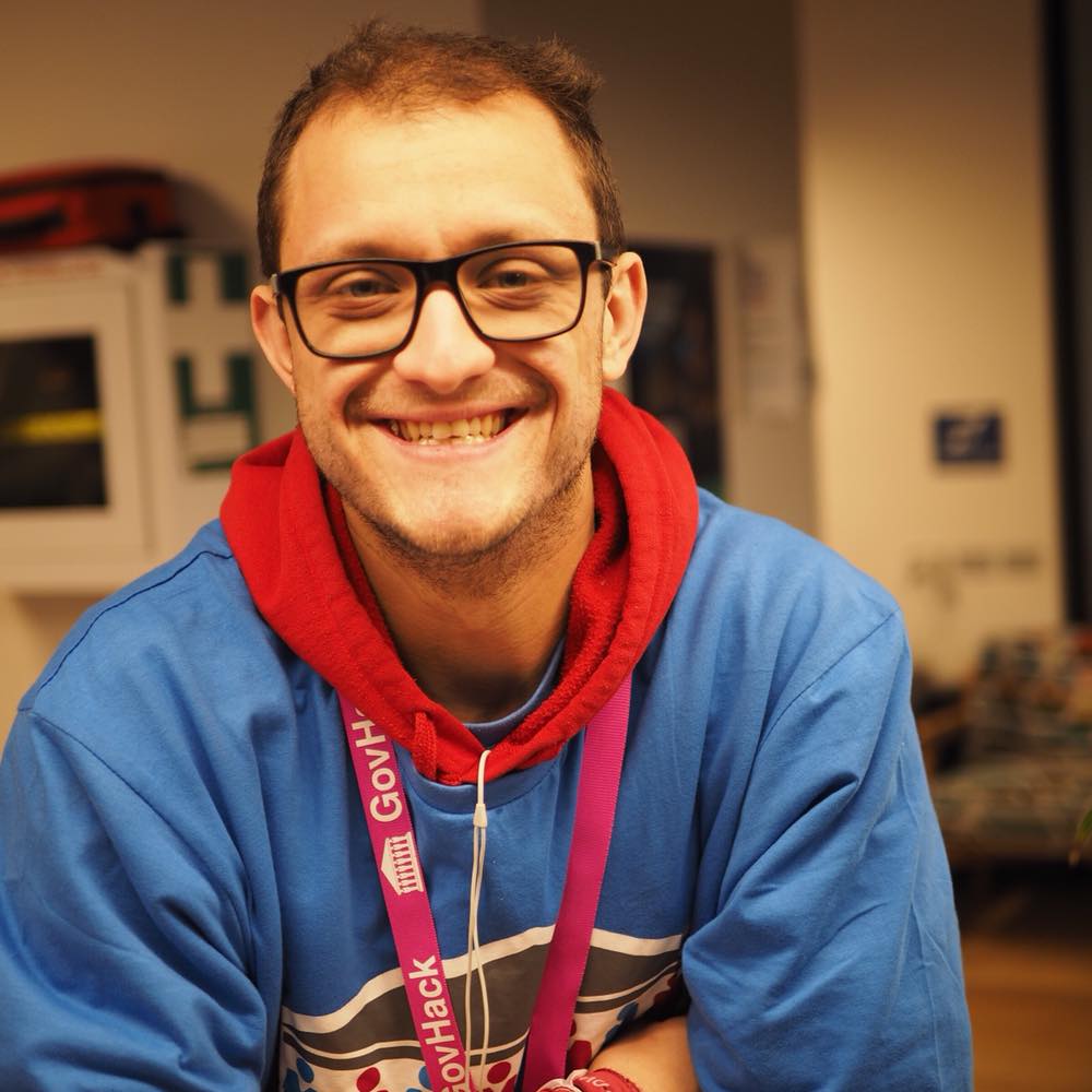 Photograph of webhost. Person is wearing glasses and a cobalt blue t-shirt with a graphic over a red jumper, with a pink lanyard that says 'GovHack'. Person is leaning on a tabletop bench.